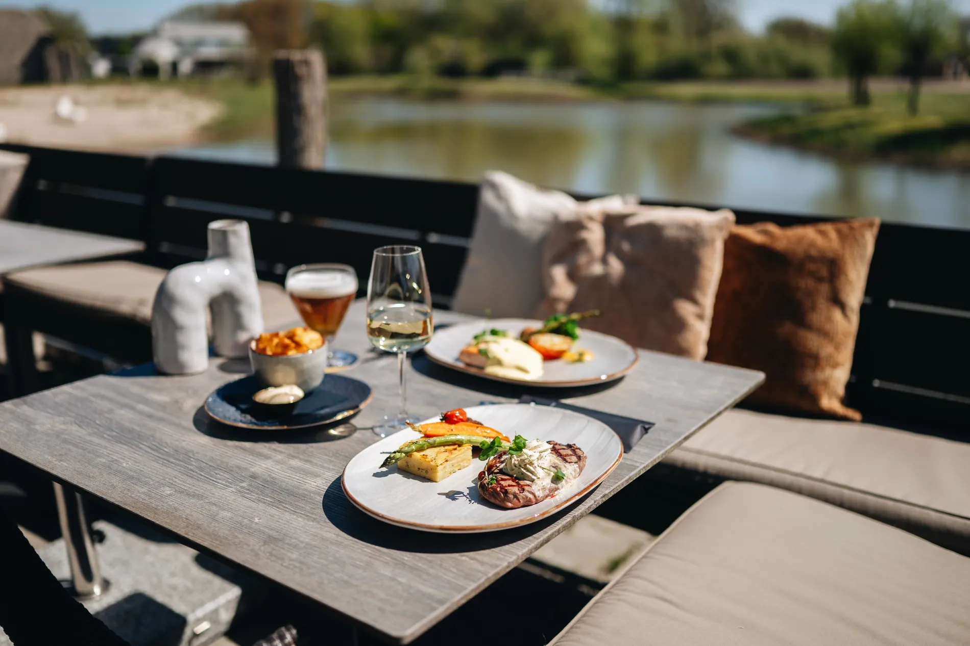 Een biefstuk met kruidenboter en een burger geserveerd op een zonnige terrastafel met kussens, direct gelegen aan een kreek of vijver op het resort.